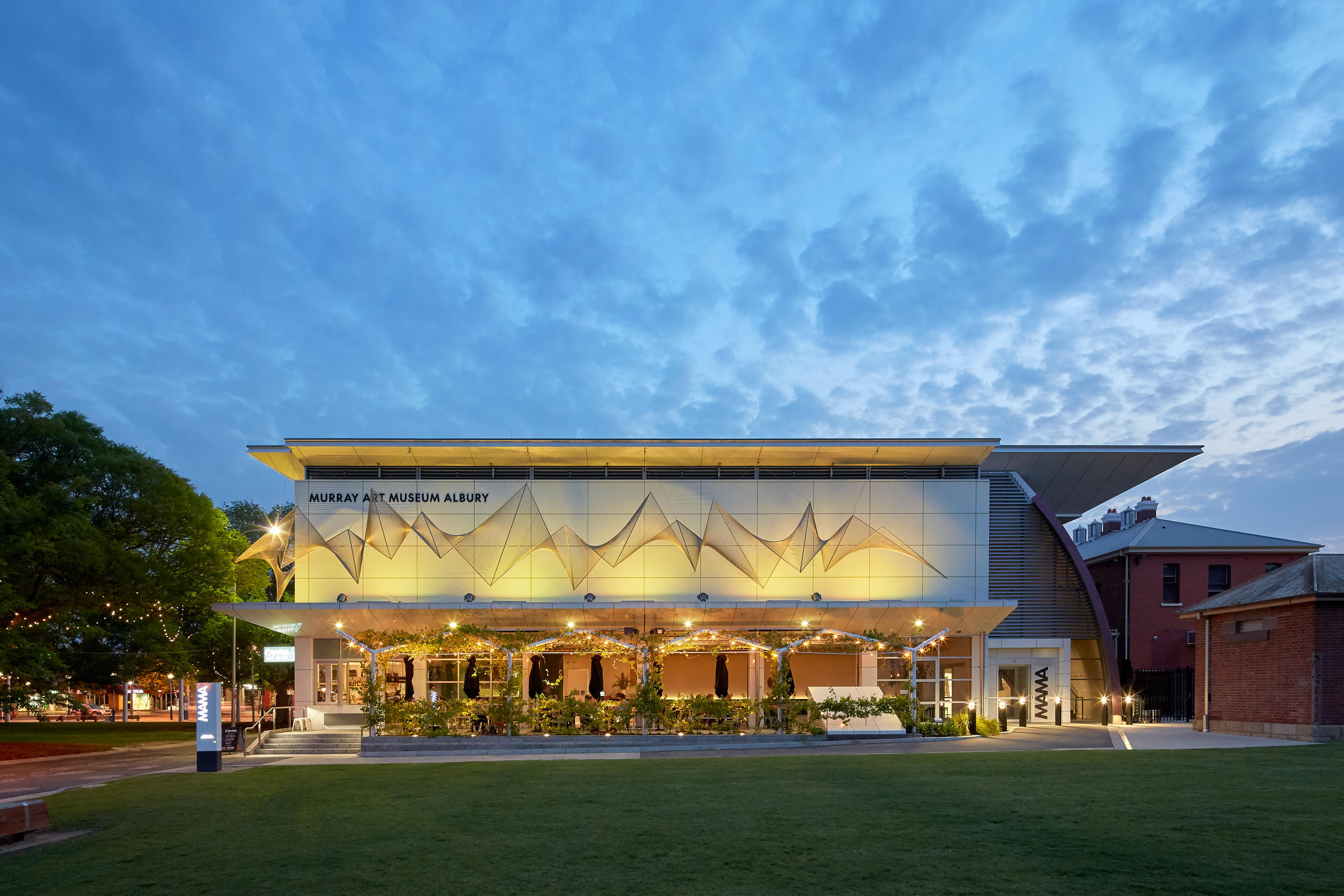 White modern rectangular building with a restaurant on ground floor, and an external wire sculpture on the second story. The building is merged with a heritage red brick building, set in an area of green lawn.